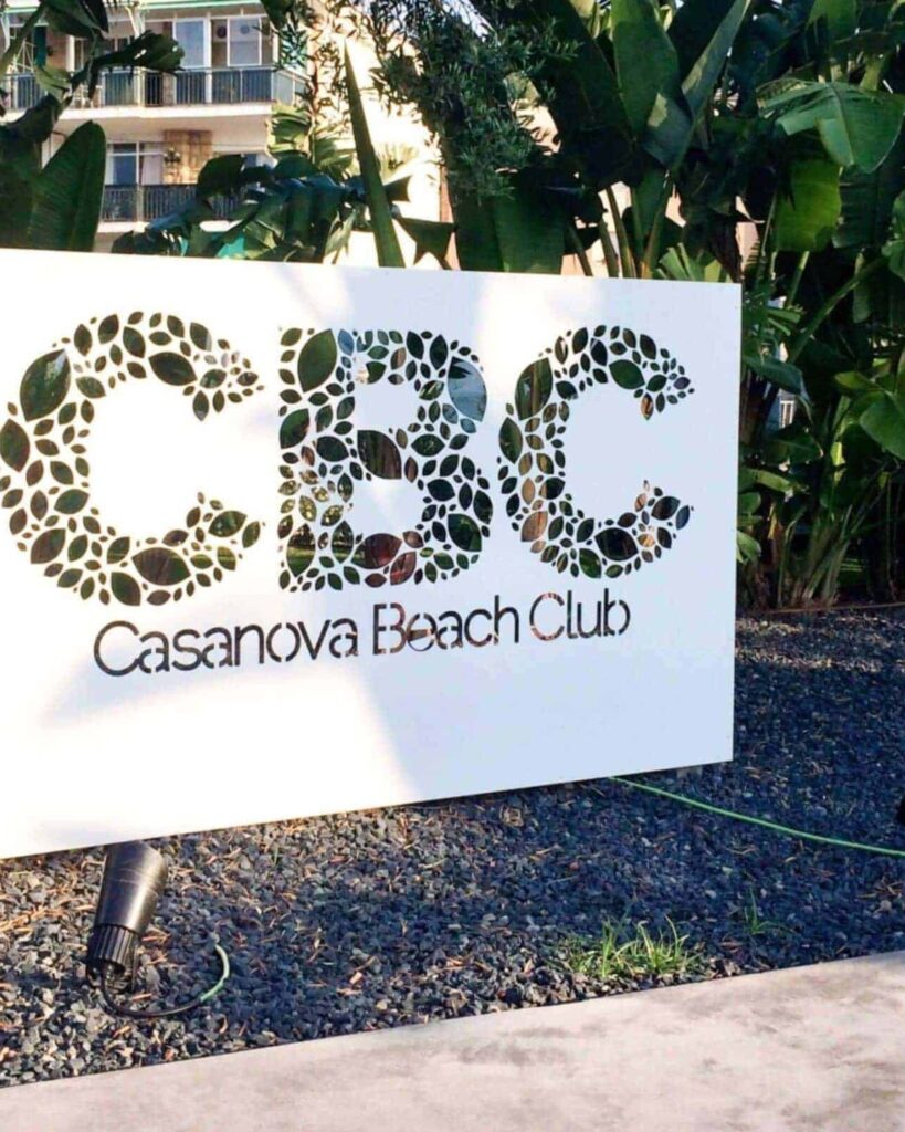 Rótulo de CBC, Casanova Beach Club, entre la vegetación del paseo marítimo de Castelldefels