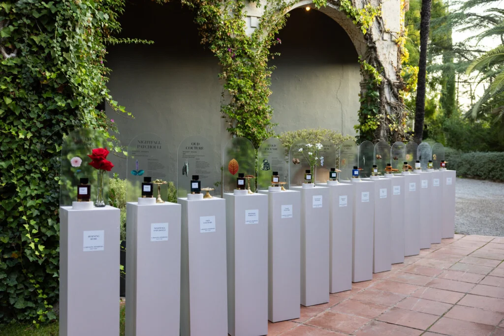 Los perfumes de Confidential en pedestales blancos bajo campanas de cristal, entre los arcos del jardín