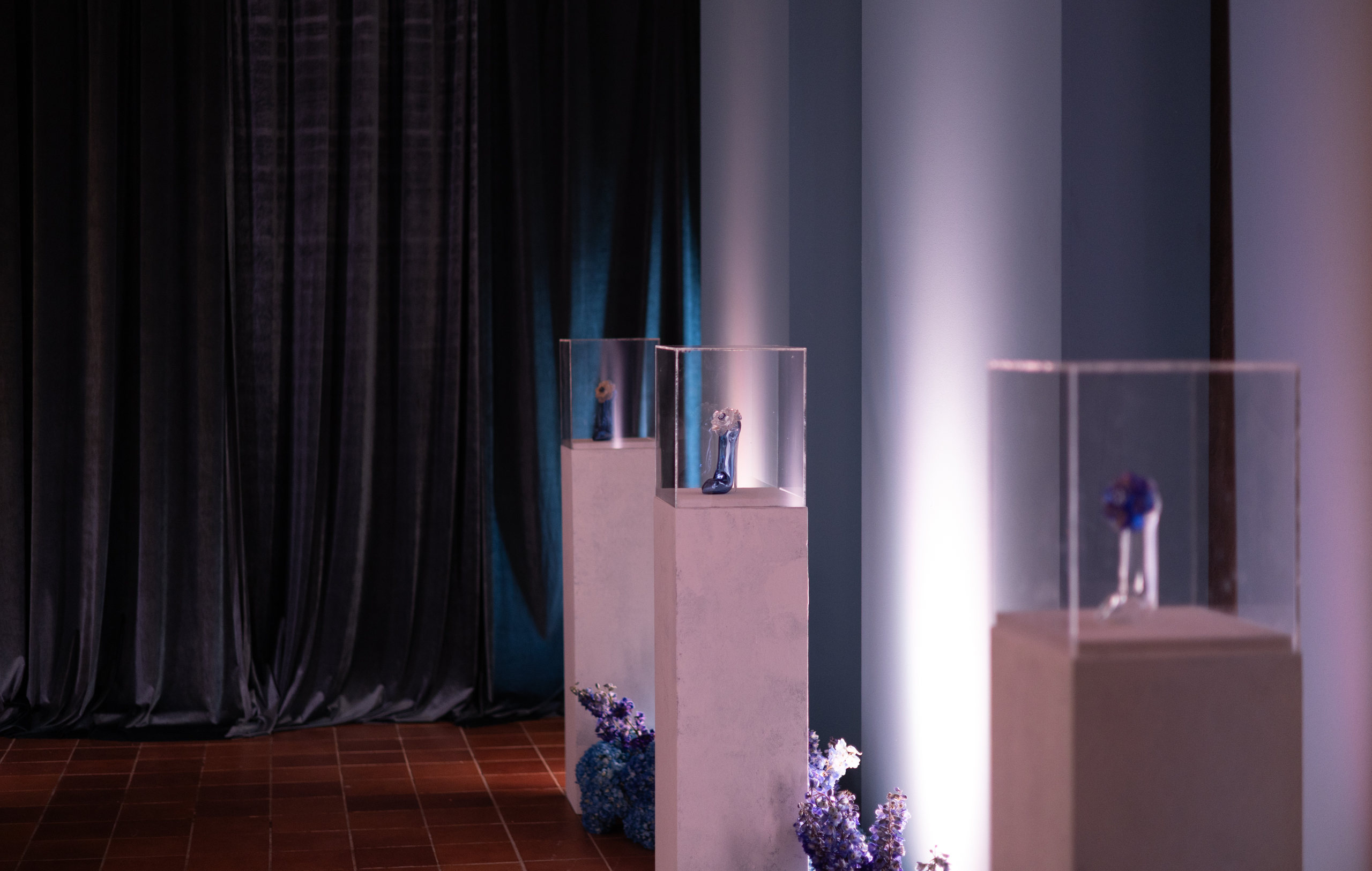 La sala a oscuras de Good Girl Murano: zapatos de vidrio en vitrinas iluminadas, entre flores lilas