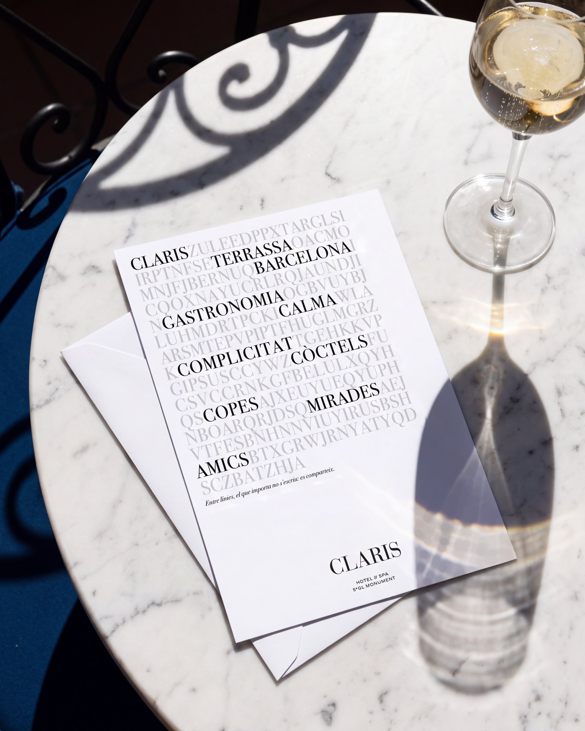 Recreación: la invitación de La Terraza del Claris 2025, con su sopa de letras, en una mesa de mármol de terraza junto a una copa de vino