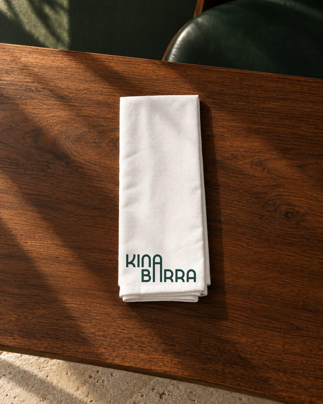 Recreación: servilleta de Kina Barra doblada, con el logotipo en verde, sobre una mesa de madera