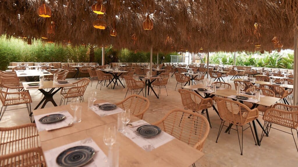 Comedor de Mamut Beach Club con mesas de madera y sillas de fibra bajo un techo vegetal