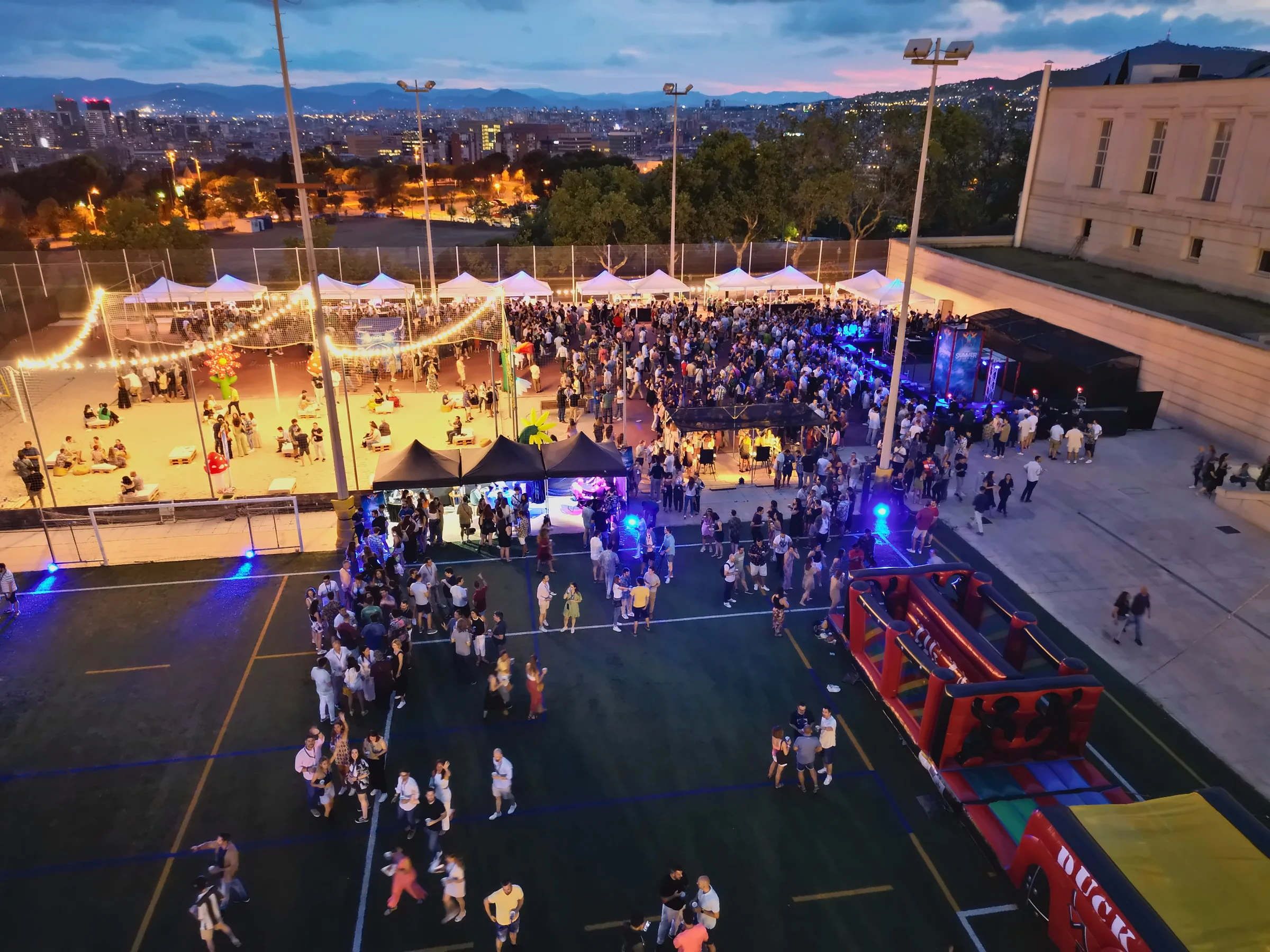 Vista aérea del Summer Sound de NTT DATA en el INEFC al anochecer: carpas, playa y escenario encendidos, con Barcelona al fondo
