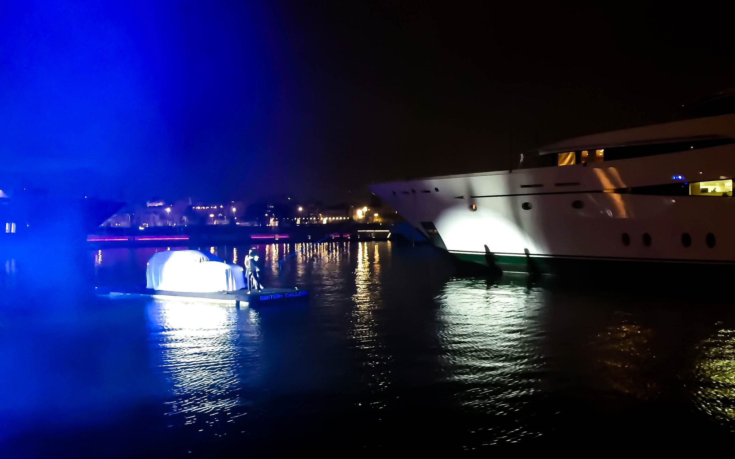 De noche, la plataforma cruza el puerto con el coche cubierto, entre un haz de luz azul y la niebla