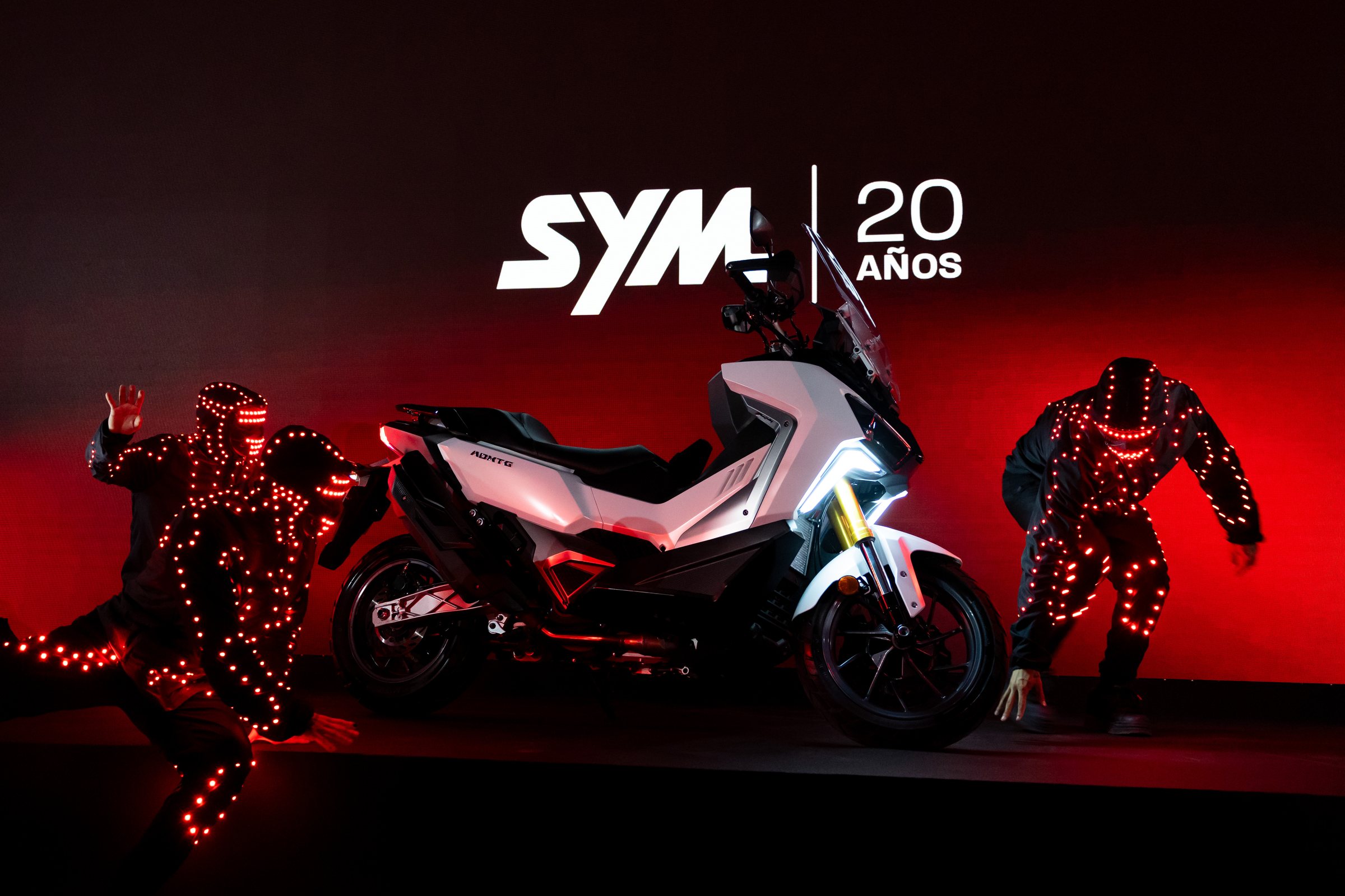 Bailarines con trajes de luz alrededor de un ADX-TG blanco ante el logotipo de SYM 20 años