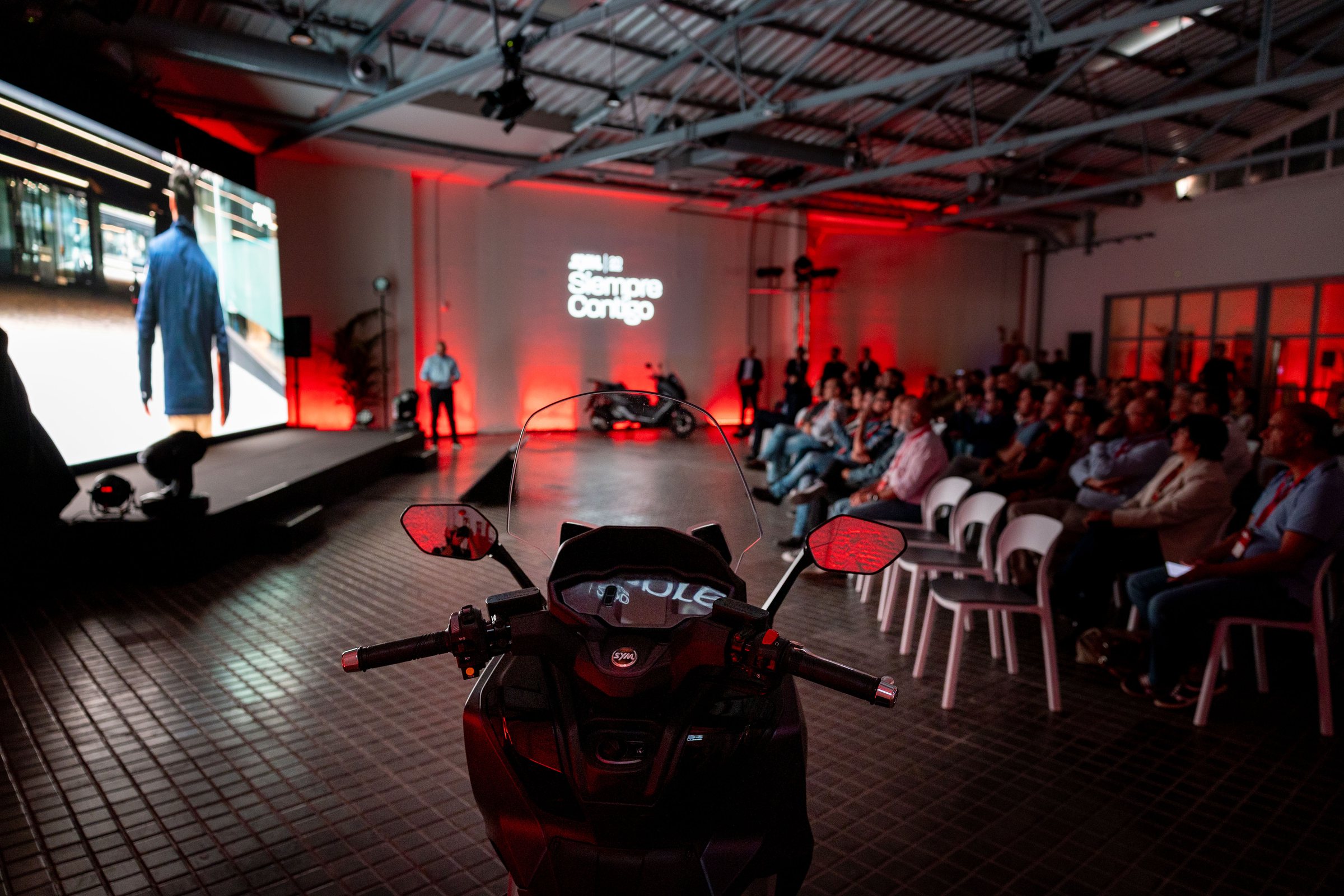 Un scooter SYM en primer plano y, al fondo, la sala del 20 aniversario con la pantalla de «Siempre Contigo»