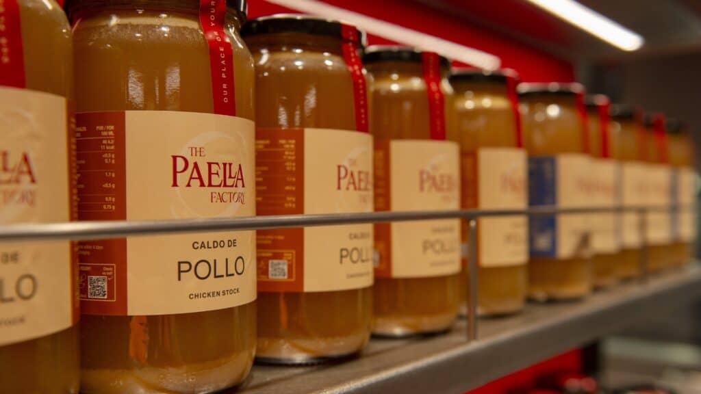 Botes de caldo de pollo de The Paella Factory con su etiqueta, en la estantería de la parada