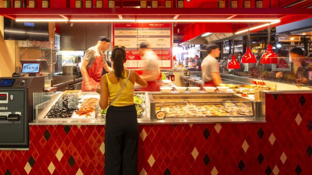 Parada de The Paella Factory en el Mercat de la Boqueria, con el mostrador rojo y el equipo cocinando