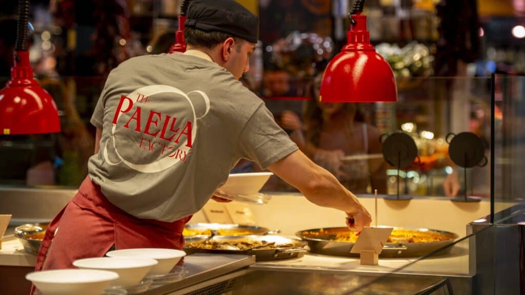Cocinero de The Paella Factory sirviendo paella con la camiseta de la marca