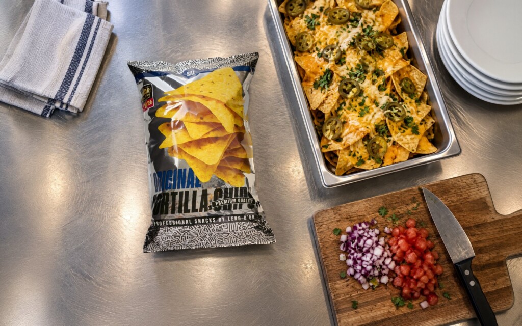 Recreación: bolsa de 450 g de Original Tortilla Chips de Leng-d’Or en la cocina de un restaurante, junto a una bandeja de nachos gratinados y la tabla con cebolla y tomate