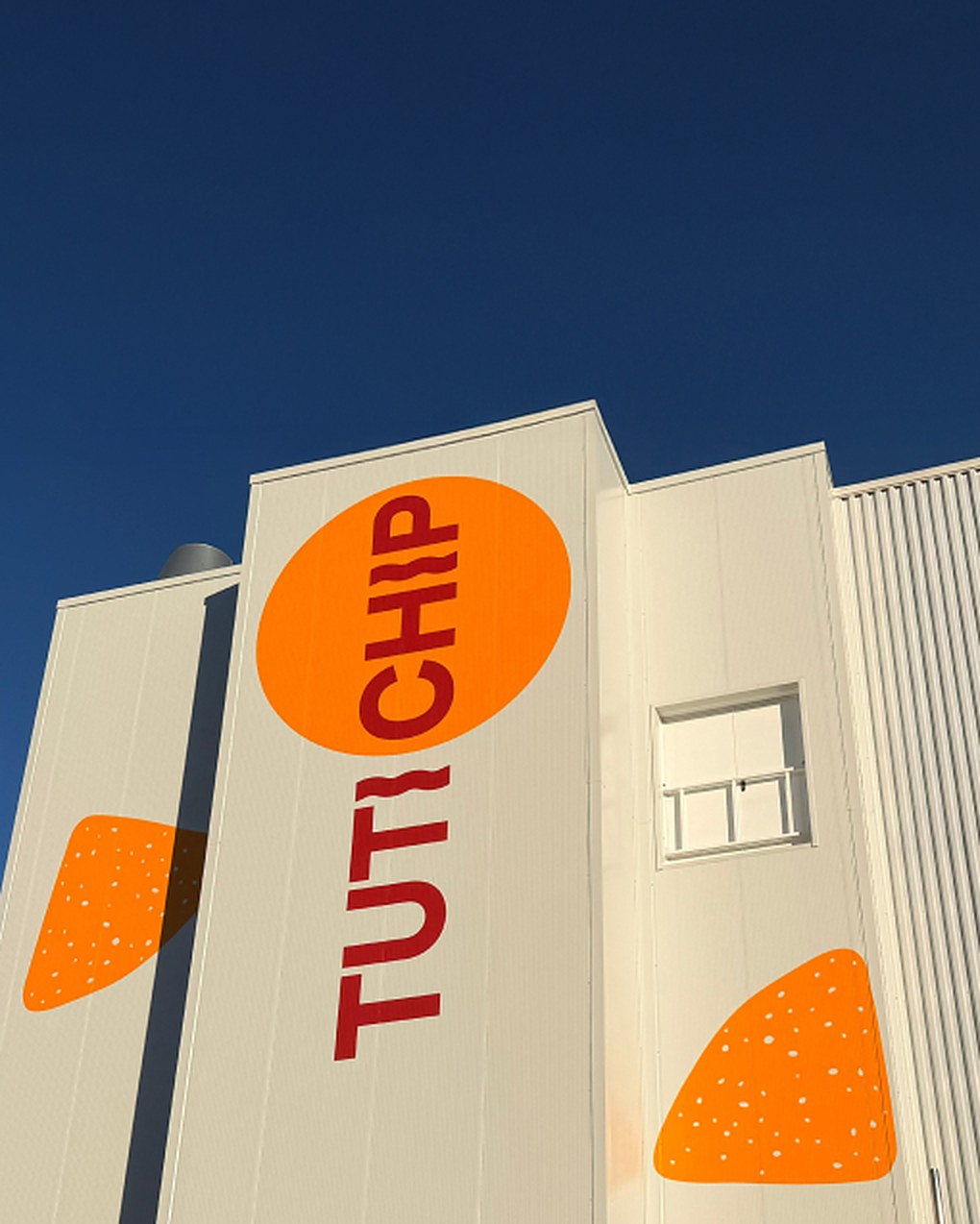La fachada de la fábrica de Tutichip, rotulada: TUTI en granate, CHIP dentro de un gran círculo naranja y dos snacks naranjas pintados