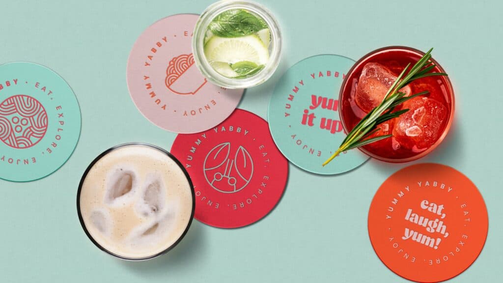 Posavasos de Yummy Yabby con iconos y lemas de la marca junto a varios cócteles