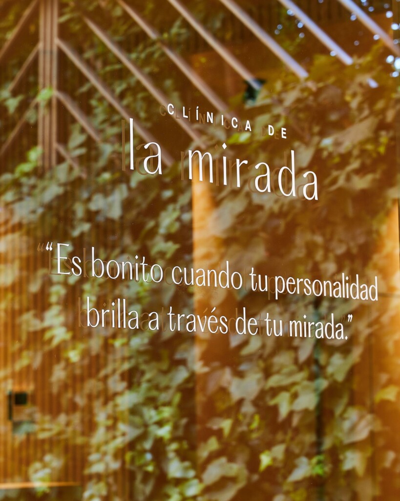 Clínica de la Mirada window vinyl, with the logo and the line “Es bonito cuando tu personalidad brilla a través de tu mirada” (It’s beautiful when your personality shines through your eyes), over ivy in the sun