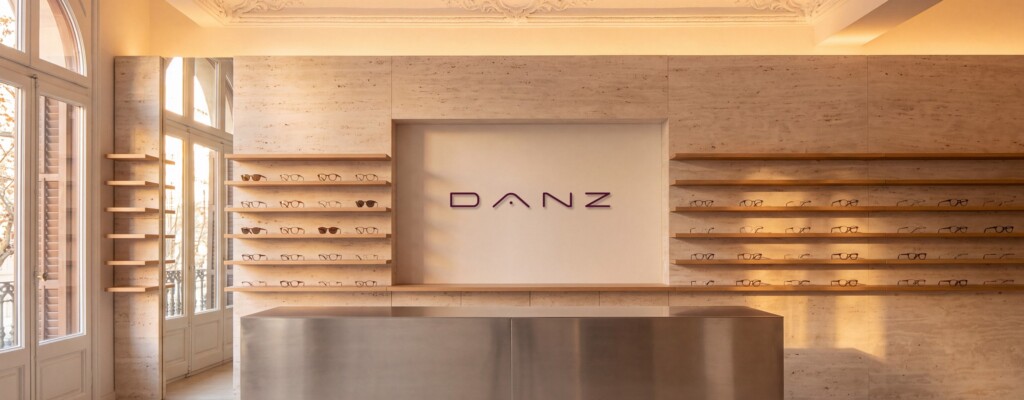 Recreación: interior de una tienda de DANZ Barcelona: muro de travertino con estantes de roble, mostrador de acero y el logotipo en burdeos bajo un techo modernista