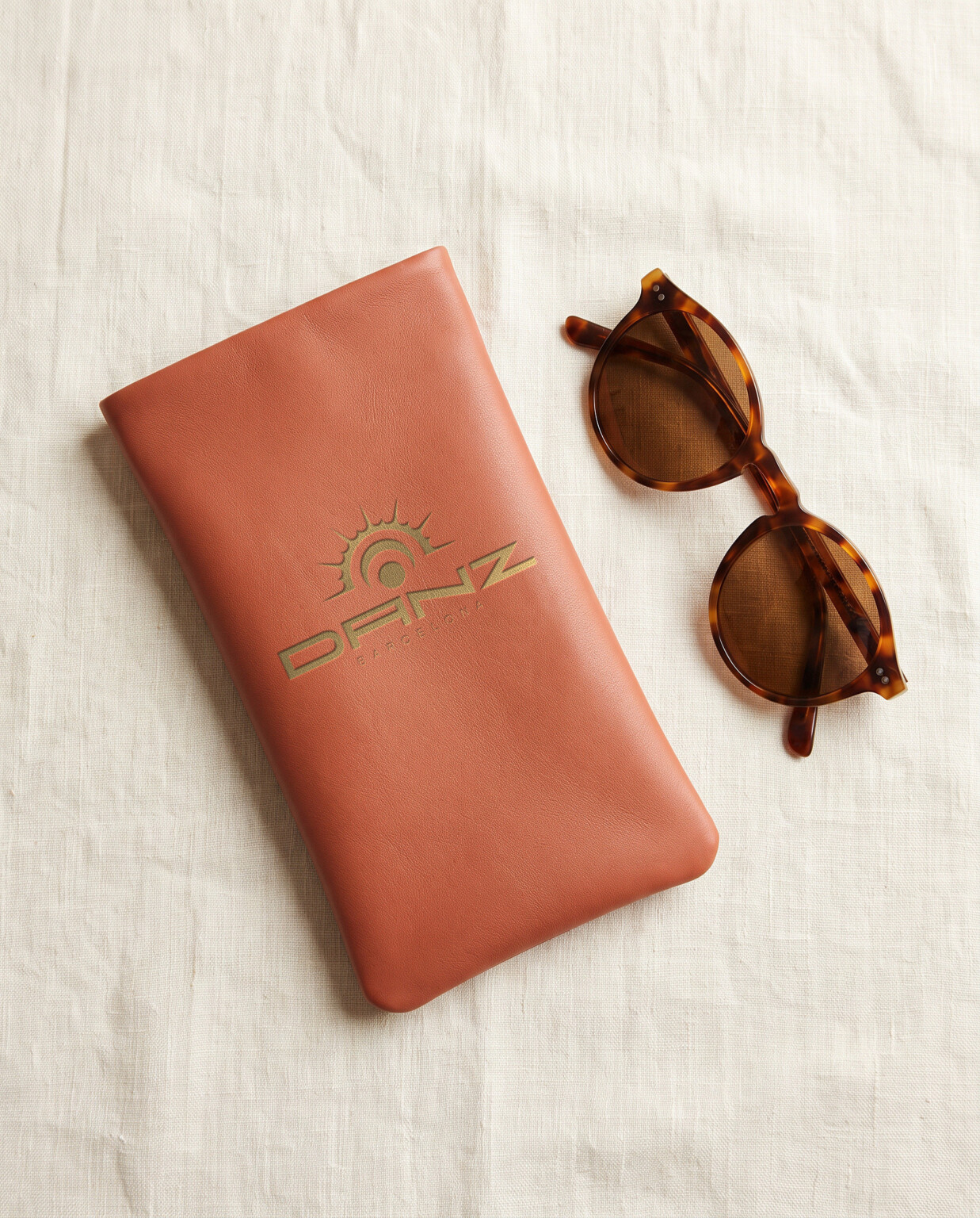 Recreación: funda de piel terracota con el logotipo del sol «Un universo cruzado» de DANZ estampado en oro, junto a unas gafas de sol