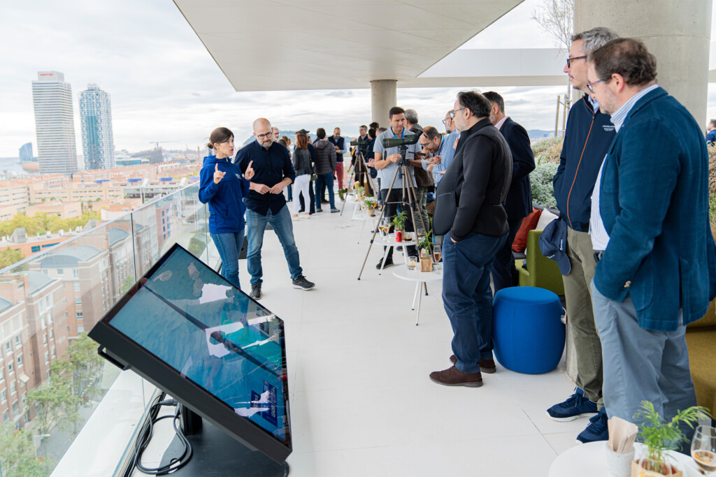Invitados de NTT DATA en la terraza frente al mar durante la Copa América de vela 2024 en Barcelona, con pantallas táctiles y telescopios