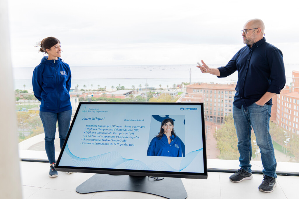 Una regatista profesional y el presentador junto a una pantalla de NTT DATA, en la terraza frente al mar