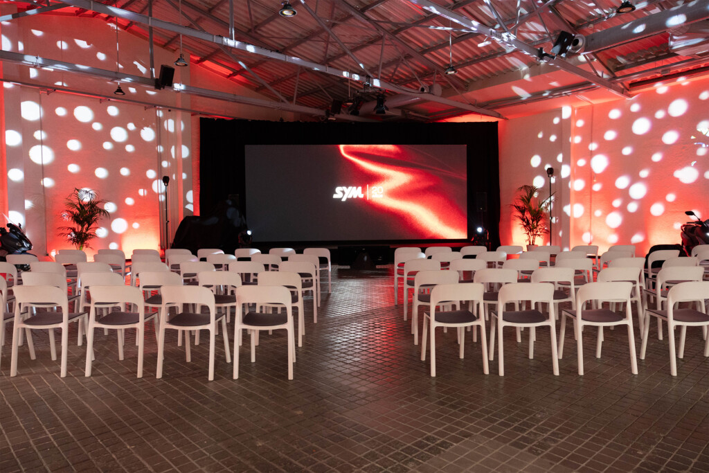 The room set for SYM España’s 20th anniversary, with white chairs, red and white light projections and the event screen
