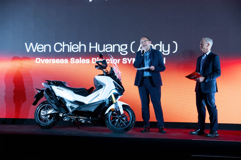 A talk on stage at SYM España’s 20th anniversary beside a SYM scooter, in front of the event screen