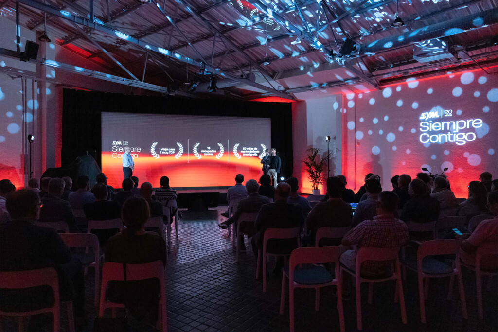 The room at SYM España’s 20th anniversary during the talks, with the laurels on screen and “Siempre contigo” (Always with you) projected on the wall